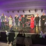 20260315_190351 Grupa osób stoi na scenie, część trzyma mikrofony. Tło jest jasne z ozdobą z kolorowych kwiatów. W tle widać instrumenty muzyczne i dekoracje świetlne.