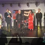 20260315_190357 Grupa sześciu osób stoi na scenie, ubranych elegancko. W tle widać perkusję i napis Dzień Kobiet. Dekoracje są kolorowe, dominują czerwone i różowe akcenty.