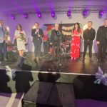 20260315_190433 Na scenie stoi siedem osób, w tym kobiety z kwiatami. Tło jest ozdobione napisami i instrumentami muzycznymi. Oświetlenie fioletowe.