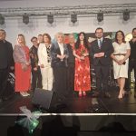 20260315_191432 Grupa trzynastu osób stoi na scenie w eleganckich strojach. Za nimi szare tło z reflektorami, z przodu stoi kilka mikrofonów. Kobieta w centrum trzyma różę i ma czerwoną sukienkę w kwiaty.