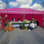 Stoisko pod różowym namiotem z napisem „Koło Gospodyń Wiejskich w GłęboCzyc”. Na stole ozdoby wielkanocne w żywych kolorach. Tło to niebo z chmurami.