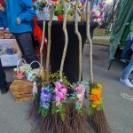 Kilka ozdobnych mioteł z kwiatami stoi przed stołem z kwiatowymi aranżacjami. Tło stanowi czerwony namiot. Obok widać ludzi w niebieskich ubraniach.