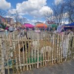 Rynek z drewnianą zagrodą dla zwierząt i kolorowymi namiotami w tle. Na niebie błękit z chmurami. Wokół ludzie spacerujący.