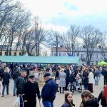 Targowisko na placu z tłumem ludzi; kolorowe namioty, drzewa bez liści, niebo z chmurami.