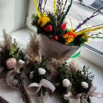 Bukiet z baziami i żółtymi kwiatami w białym koszyku stoi na parapecie. Obok cztery wiązanki z zielonymi gałązkami i koronkowymi wstążkami. Tło to widok ulicy za oknem.