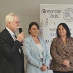 Trzy osoby stoją przed banerem Słoneczne Zioła. Jedna osoba mówi do mikrofonu. Tło jest jasne, widać flagę Polski.