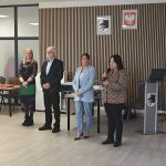 Czworo ludzi stoi na tle drewnianej ściany z herbami i białym orłem. Kobieta trzyma mikrofon, a inna zieloną teczkę. Tło ma jasne podłogi i meble.