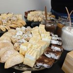 Stół z ciastkami różnych kształtów na szklanych paterach. Dominują kolory jasnobrązowy i biały. W tle słoiki z cukrem i innymi dodatkami.