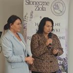Dwie kobiety stoją przed banerem „Słoneczne Zioła”. Jedna w jasnym garniturze, druga w kwiecistej bluzce rozmawia przez mikrofon. W tle komputer i jasne tło.