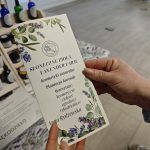 Plakat w dłoniach przedstawia ofertę farmy lawendowej. Dominują kolory fioletowy i zielony. W tle są butelki i drewniana półka.