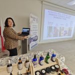 Osoba przy laptopie prezentuje slajd o kosmetykach naturalnych. Na stole stoją kolorowe butelki, w tle są flagi i plakat. Sala ma jasne ściany i podłogę.