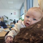 Na pierwszym planie niemowlę na ramieniu kobiety, w tle mężczyzna przy stole. Dominują jasne i pastelowe kolory. Pomieszczenie biurowe z roślinami i oknami.