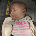 Na zdjęciu śpiące niemowlę w foteliku samochodowym. Ubrane w beżową bluzę z różowym napisem. Tło w odcieniach szarości i beżu.