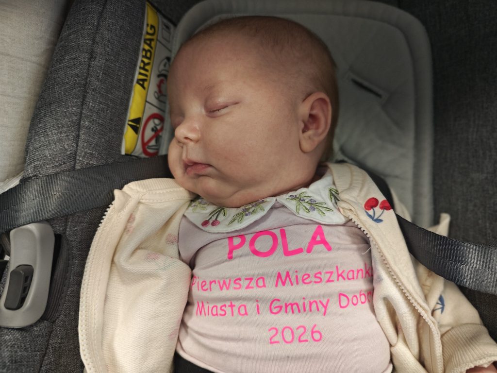 Małe dziecko śpi w foteliku samochodowym z szarymi pasami. Ubrane jest w różowe body z napisami i jasny sweterek. Tło ma szary odcień.