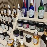 Półki z naturalnymi kosmetykami w kolorowych butelkach. Przedmioty ustawione na drewnianych stojakach. Tło w jasnych barwach.