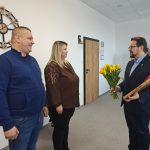 Trzy osoby w biurze: mężczyzna wręcza bukiet żółtych tulipanów parze. Na ścianie duży zegar. Eleganckie wnętrze.