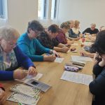 Grupa seniorów pisze przy drewnianym stole w jasnym pokoju. Mają kolorowe ubrania i trzymają kartki oraz długopisy. W tle widać okna i białe torby.