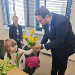 Dziewczynka siedzi na zielonym fotelu, trzymając papierową torbę. Mężczyzna w garniturze wręcza jej żółte tulipany. W tle stoi uśmiechnięty chłopiec przy oknie z żaluzjami.