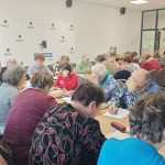 Grupa ludzi siedzi przy dużym stole, pisząc na kartkach. Pomieszczenie ma jasne ściany i szklane drzwi. Logo i napisy w tle.