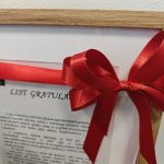 Certyfikat w drewnianej ramce z czerwoną wstążką i kokardą. Tło jasne, gładkie. Widoczny napis LIST GRATULACYJNY.