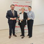 652294867_951497793892409_4732967781104302922_n Trzy osoby w formalnym stroju stoją przed banerem „Mazowsze”. Środkowa osoba trzyma dokument. Tło jest jasne, podłoga z drewna.