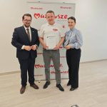 652581068_951498343892354_2297855323498971524_n Trzy osoby stoją przed białym tłem z czerwonymi napisami, środkowa osoba trzyma dokument. Scena odbywa się w jasnym pomieszczeniu.