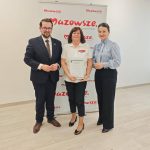 652980291_951497993892389_7621754299551251040_n Trzy osoby stoją przed białym tle z logo Mazowsze. Kobieta w środku trzyma dyplom. Podłoga jest jasnobrązowa.