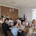 653021616_951497550559100_8795195196125543519_n Spotkanie biznesowe przy drewnianym stole, 10 osób w eleganckich ubraniach. Kobieta prezentuje na tle slajdu z białą ścianą, obok flagi. Pomieszczenie z drewnianą ścianą i sufit z białymi panelami.