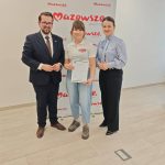 653047723_951499263892262_5428796838585882107_n Trzy osoby stoją na jasnym tle z logo Mazowsze. Osoba w środku trzyma certyfikat. Tło jest białe z czerwonymi napisami.