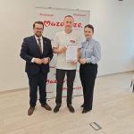 653049115_951498050559050_2612671153124182874_n Na zdjęciu są trzy osoby stojące na tle baneru Mazowsza. Osoba w środku trzyma dyplom. Podłoga jest w jasnym, drewnianym odcieniu.