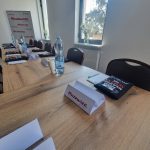 653051615_951496090559246_969396108390680505_n Długi stół z drewnianym blatem, na którym znajdują się notatniki i butelki wody. Na stole leżą wizytówki z napisem Mazowsze. W tle znajduje się jasna ściana z logo i okno z widokiem na zieleń.