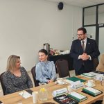 653056345_951496583892530_2915171223135052568_n Grupa ludzi siedzi przy stole w sali konferencyjnej. Mężczyzna w ciemnym garniturze stoi, reszta rozmawia. Na stole są filiżanki, notatki i dzbanki.