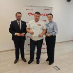 653123995_951498150559040_3695347357427231775_n Trzy osoby stoją na drewnianej podłodze, na tle baneru z napisem Mazowsze. Osoba w środku trzyma certyfikat. Ubrania w tonacji bieli, granatu i czerni.