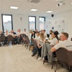 653420944_951497613892427_6501654154381455049_n Grupa ludzi w białych koszulkach siedzi przy stołach w jasnym pomieszczeniu. Na ścianie wisi krzyż i baner z logotypem. Tło jest białe z dużymi oknami po lewej stronie.