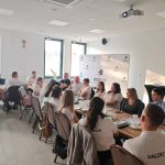 653454344_951496157225906_7485938921460931033_n Grupa osób siedzi przy stole w jasnym pokoju. Ściana z logo i białe koszulki uczestników. W tle krzyż i duże okna.