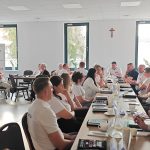 653519531_951498757225646_6065964537427309782_n Spotkanie w jasnym pokoju z dużymi oknami; ludzie siedzą wokół stołu, niektórzy w białych koszulkach. W tle widoczny banner Mazowsze; na ścianie krzyż. Na stole notatniki, filiżanki i dzbanki z