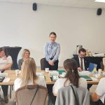 653640930_951496747225847_7606648257531855487_n Grupa osób siedzi przy stole na spotkaniu. Na stole widać notatki i kubki, w tle stół z kawą i herbatą. Ściany są białe, a światło jasne.