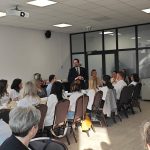 653649449_951497500559105_3333735202462331704_n Grupa ludzi siedzi przy stole konferencyjnym, mężczyzna w garniturze przemawia. Jasne ściany, z projekcją na suficie. Tło biurowe, szyby i światło słoneczne.