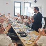 653700174_951495667225955_6538582003472237269_n Na zdjęciu grupa ludzi siedzi przy stole w jasnym pomieszczeniu. Wszyscy patrzą na stojącego mężczyznę w garniturze, klaskając. W tle widoczne są okna oraz krzyż na ścianie.