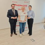 653700260_951506527224869_6901310090542824366_n Trzy osoby stoją na tle białego banera z czerwonym napisem Mazowsze. Środkowa osoba trzyma dyplom. Podłoga jasnobrązowa.