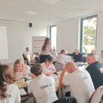 653701886_951496423892546_355938241869893845_n Grupa ludzi w białych koszulkach siedzi przy stołach w jasnej sali. W tle widoczne są okna i ekran z napisem Mazowsze. Pomieszczenie jest przestronne i dobrze oświetlone.