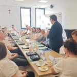 653702272_951497273892461_5880025853723032917_n Grupa osób siedzi przy długim stole w sali konferencyjnej. Mężczyzna w garniturze stoi i mówi do uczestników. Tło jasne, okna i krzyż na ścianie.