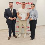 653703284_951498717225650_2405420444149939719_n Trzy osoby stoją przed białą tablicą z logo Mazowsze. Osoba w środku trzyma certyfikat. Podłoga jest drewniana.