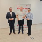 653703873_951496950559160_5285223824572229636_n Trzy osoby stoją na tle białej ścianki z czerwonym napisem Mazowsze. Mężczyzna w środku trzyma certyfikat. Podłoga jest jasna, drewniana.