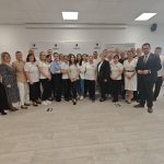 653743344_951506577224864_7952819342474301773_n Grupa ludzi pozuje do zdjęcia w jasnym pomieszczeniu z drewnianą podłogą. Większość osób ubrana jest w białe koszulki. W tle widoczna jest szara ściana z logo.