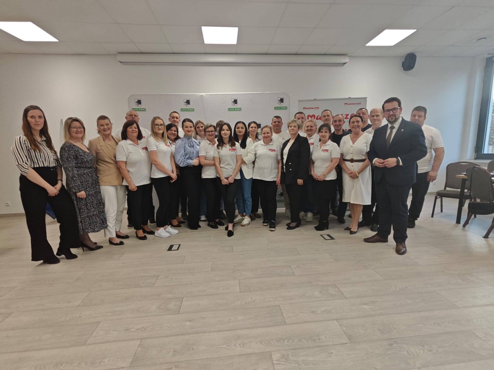 Grupa ludzi pozuje do zdjęcia w jasnym pomieszczeniu z drewnianą podłogą. Większość osób ubrana jest w białe koszulki. W tle widoczna jest szara ściana z logo.