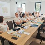 654207855_951498983892290_8191548791620465069_n Grupa ludzi w białych koszulkach siedzi przy stole z notatnikami. Na stole filiżanki kawy i dzbanki z wodą. W tle widać prezentację na ścianie.