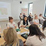 654235261_951499027225619_2651248317081899519_n Grupa ludzi siedzi przy stole w sali konferencyjnej. W tle widać prezentację na ekranie i plakaty z napisem Mazowsze. Uczestnicy mają na sobie białe koszulki z imionami.