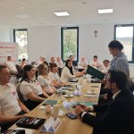 654346374_951496520559203_3955513351514517019_n Grupa ludzi siedzi przy stole z dokumentami, w jasnym pomieszczeniu z białymi ścianami. Osoba z przodu trzyma zieloną teczkę. W tle logo Mazowsze na białym tle.