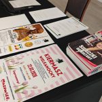 654358417_951499170558938_4364365859269971453_n Na czarnym stole leżą kolorowe ulotki i magazyny. Ulotki promują wydarzenia, jak Kiermasz Wielkanocny. W tle widoczne są krzesła i podłoga.